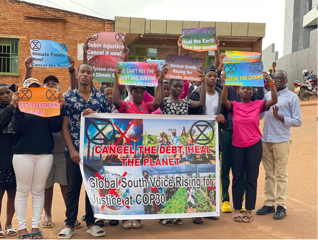 Ein Foto einer Gruppe, die vor einem Backsteinhaus steht und Schilderhält. Auf den Schildern sind XR-Symbole und Slogans zu sehen, auf demgrössten steht "cancel the debt heal theplanet".