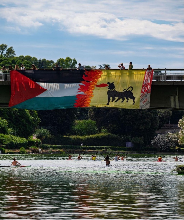 A photo of a bridge over a river. There are people on the bridge holding a very large banner over the side, with the Palestinian flag, the other side is yellow with a black animal on it.