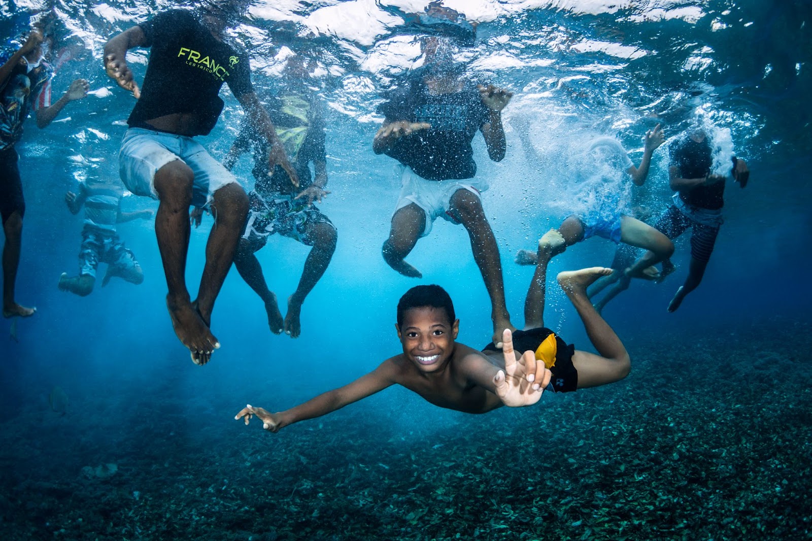 Boys
swimming. Da sotto l'acqua, vediamo otto ragazzi che nuotano. Sette di loro hanno latesta fuori dall'acqua e non possiamo vederne il volto, ma un ragazzo hanuotato sott'acqua e sta galleggiando orizzontalmente, sorridendo eindicando versol'alto.