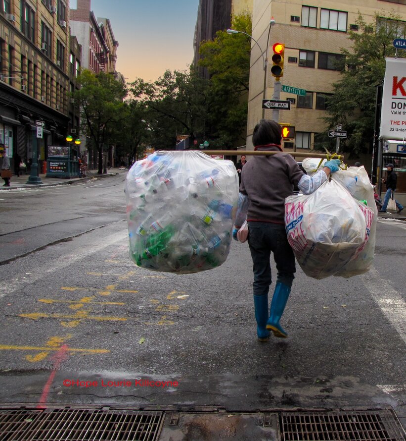 Eine Person überquert bei Einbruch der Dunkelheit die Straße in New YorkCity. Sie trägt eine Holzstange über der rechten Schulter, an deren Endenzwei riesige durchsichtige weiße Plastiktüten mit leeren Plastikflaschenbalancieren.